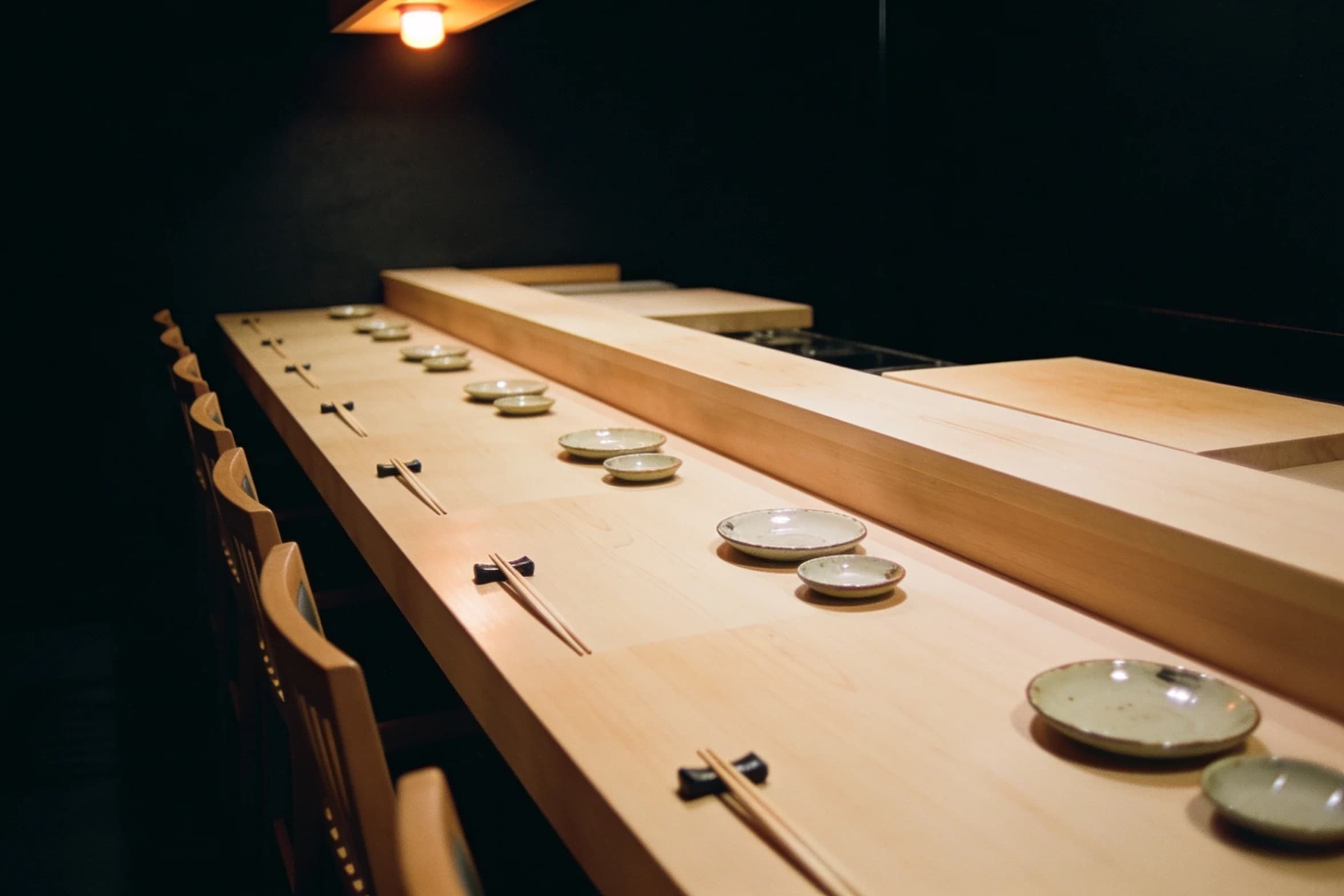 An empty hinoki sushi counter in Ginza, eight seats set with chopstick rests under a single warm light.