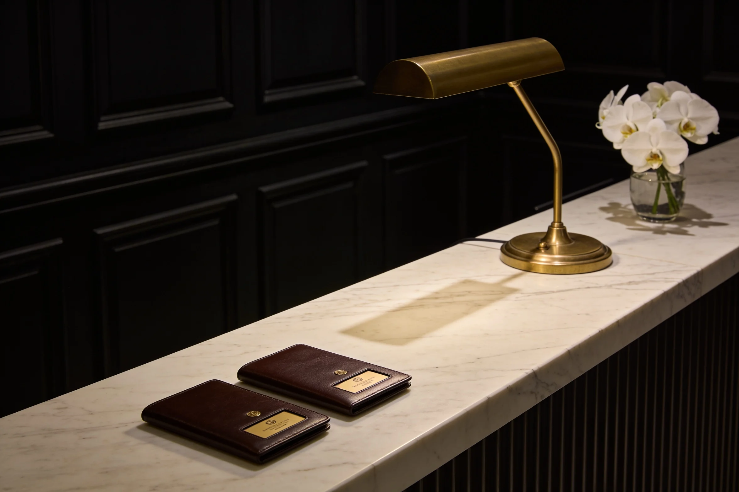 A hotel reception desk late at night, two key card wallets on marble under a low brass lamp.