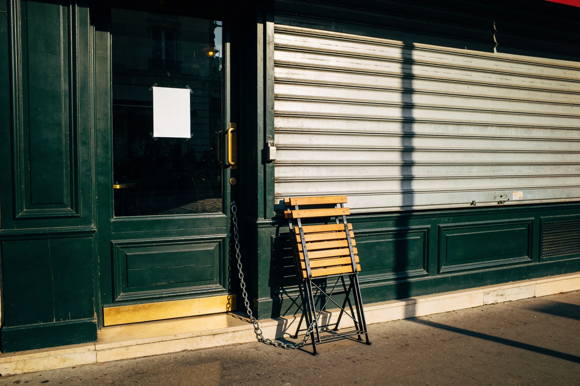 A shuttered Paris restaurant front in summer sun, a plain paper notice taped inside the glass door and a folded cafe chair chained beside it.
