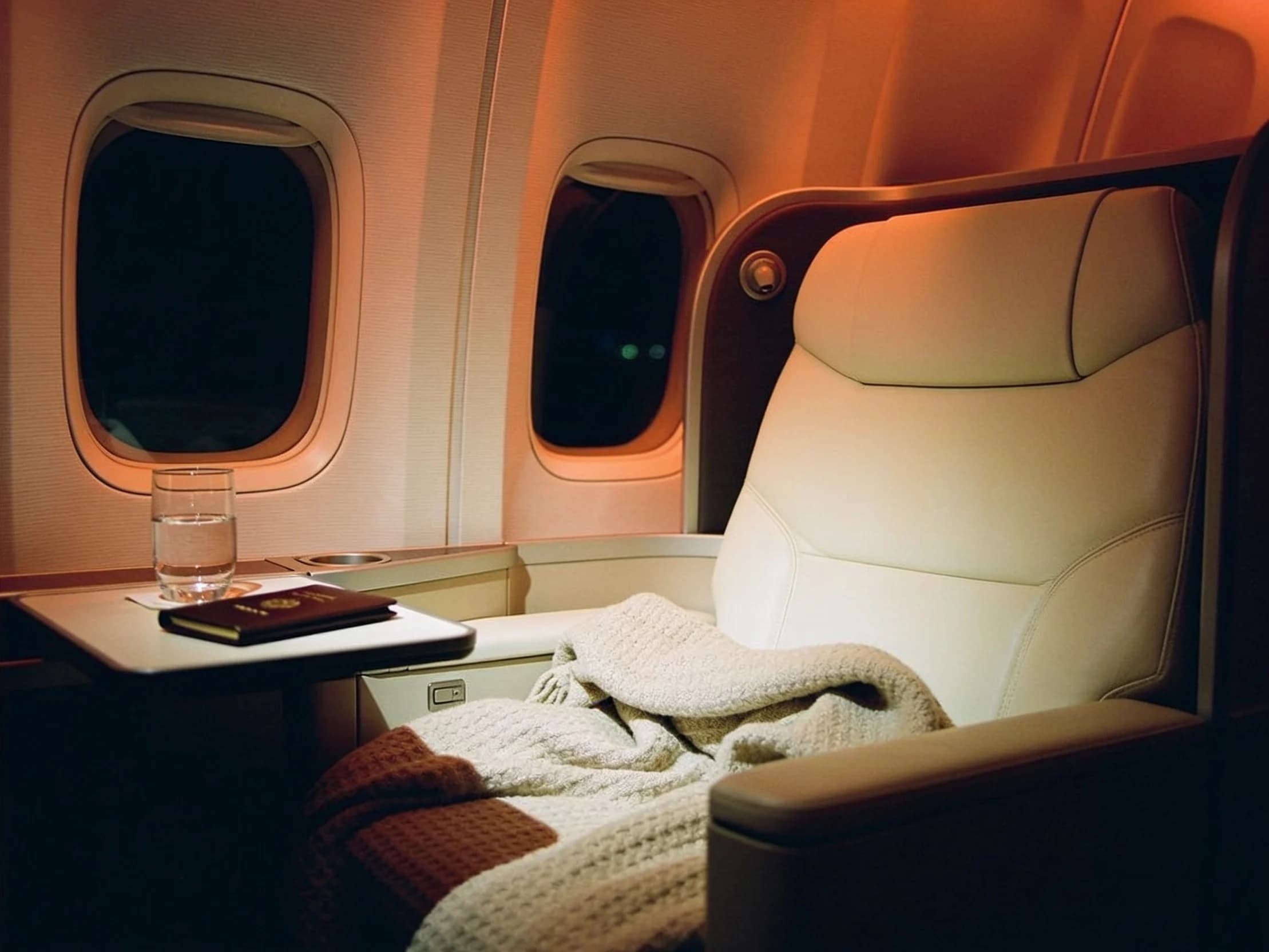A first class airline suite at night, a cashmere blanket and a leather passport wallet under amber light.