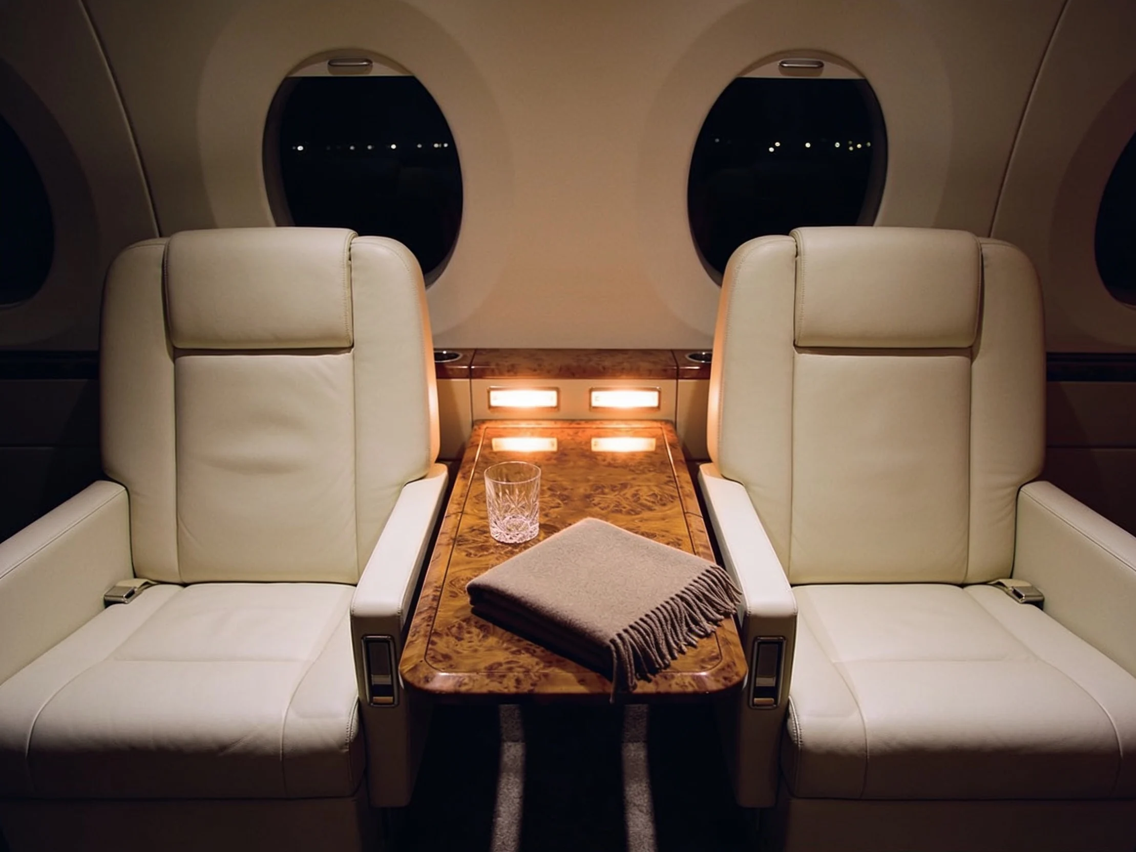 The cabin of a private jet at night, two cream leather seats facing across a walnut table.