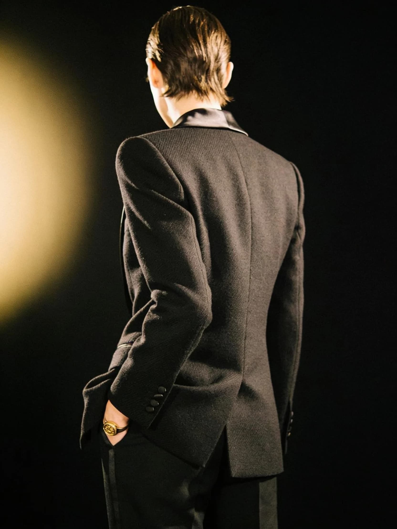 Amara in a sharply tailored black tuxedo, photographed from behind against a dark ground.