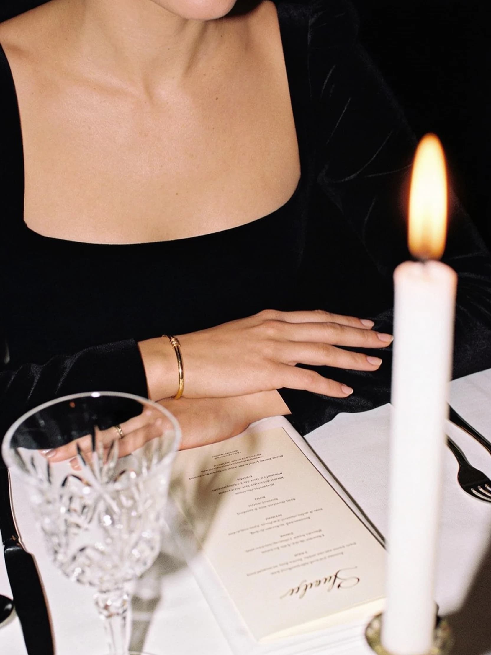 Charlotte's hands resting on a white linen dinner table beside a crystal glass, by candlelight.