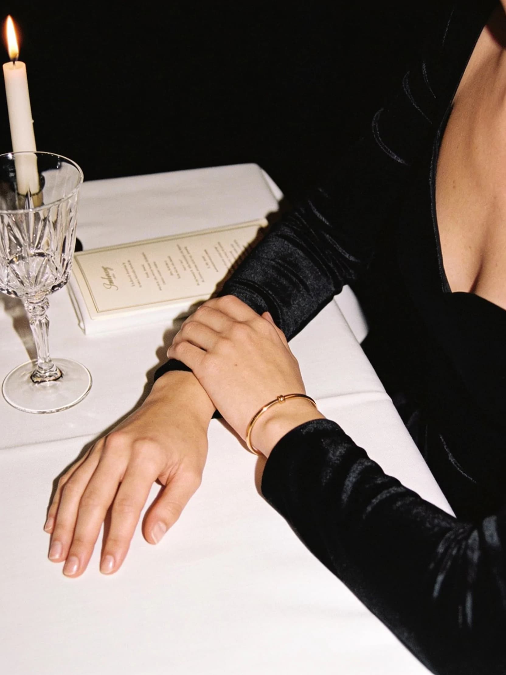 Élodie's hands resting on a white linen dinner table beside a crystal glass, by candlelight.