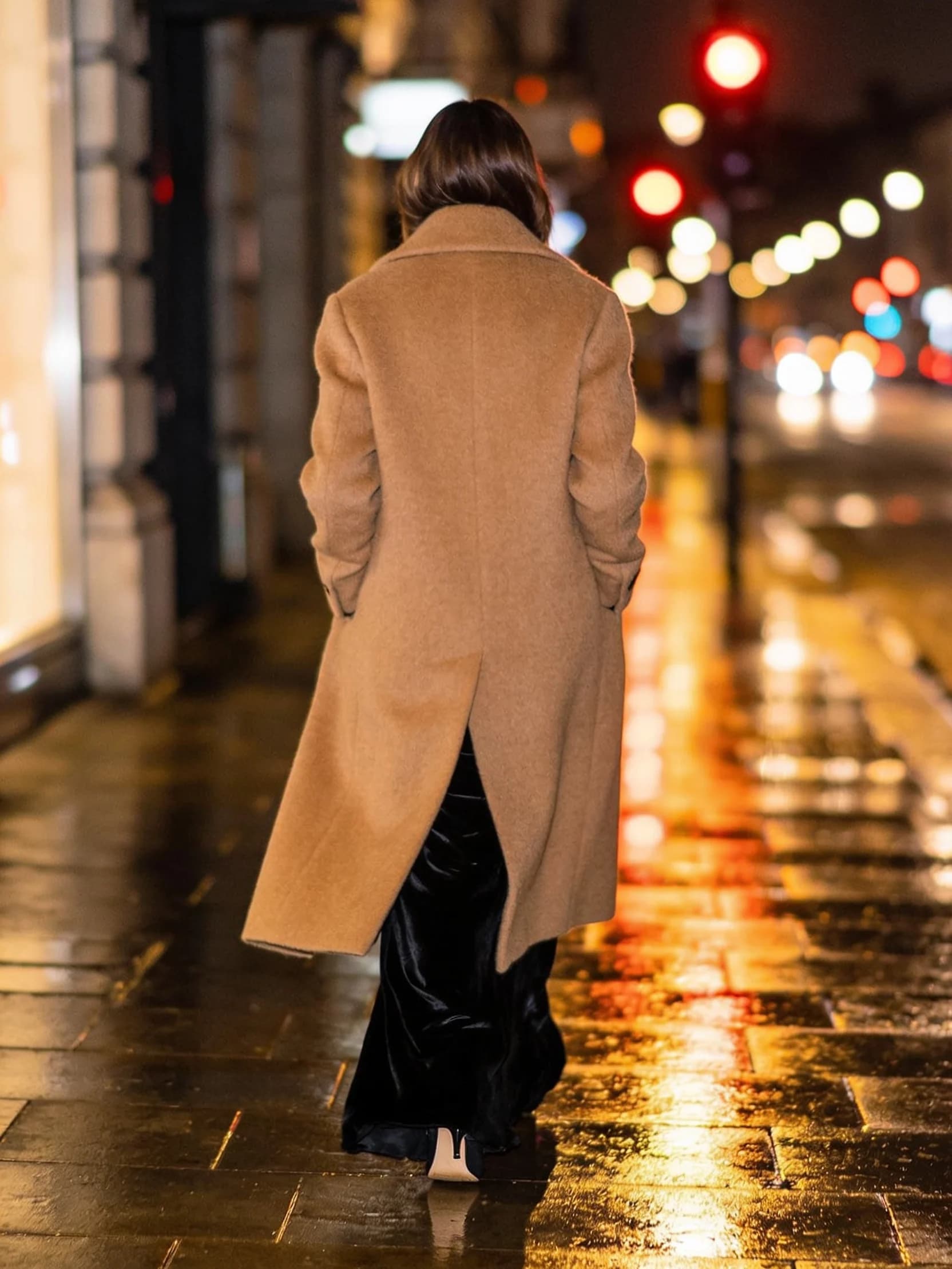 Kanya in a long overcoat, walking away along a rain-wet city street at night.