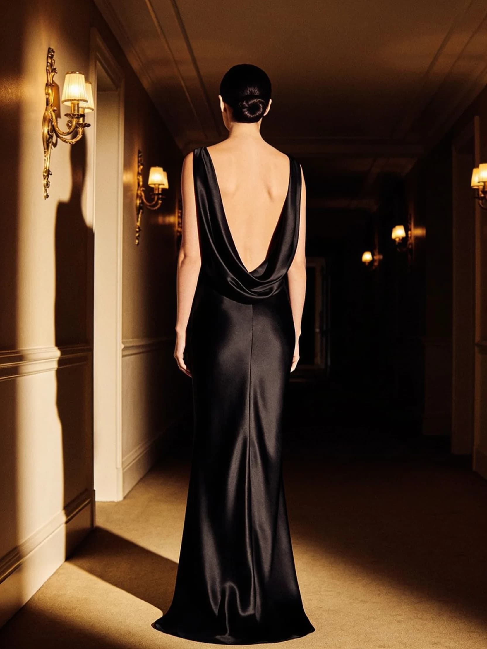 Layla, photographed from behind in a floor-length black silk gown in a lamplit hotel corridor.