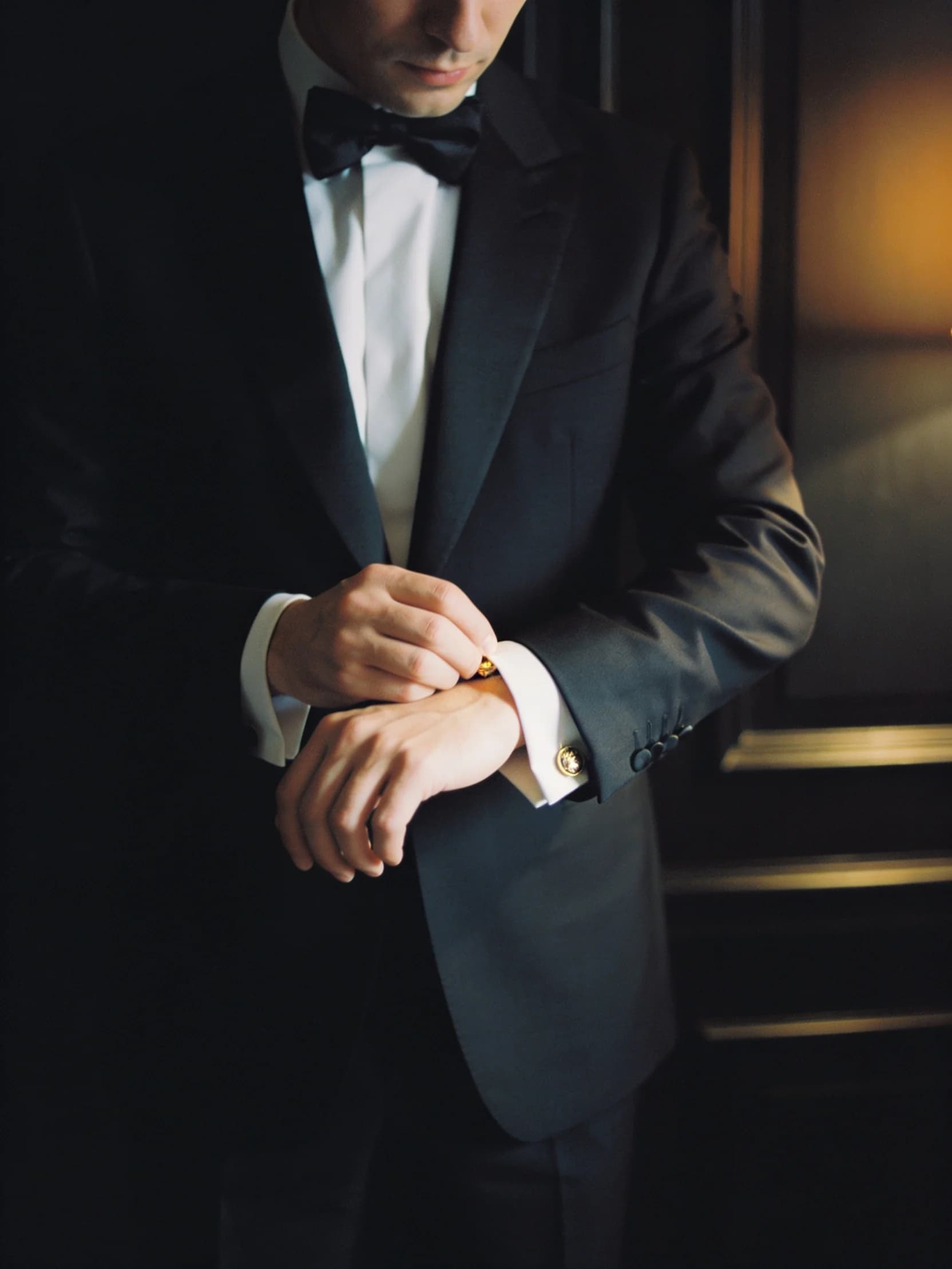 Marcus in a black dinner jacket, adjusting a cuff, photographed from behind in a panelled room.
