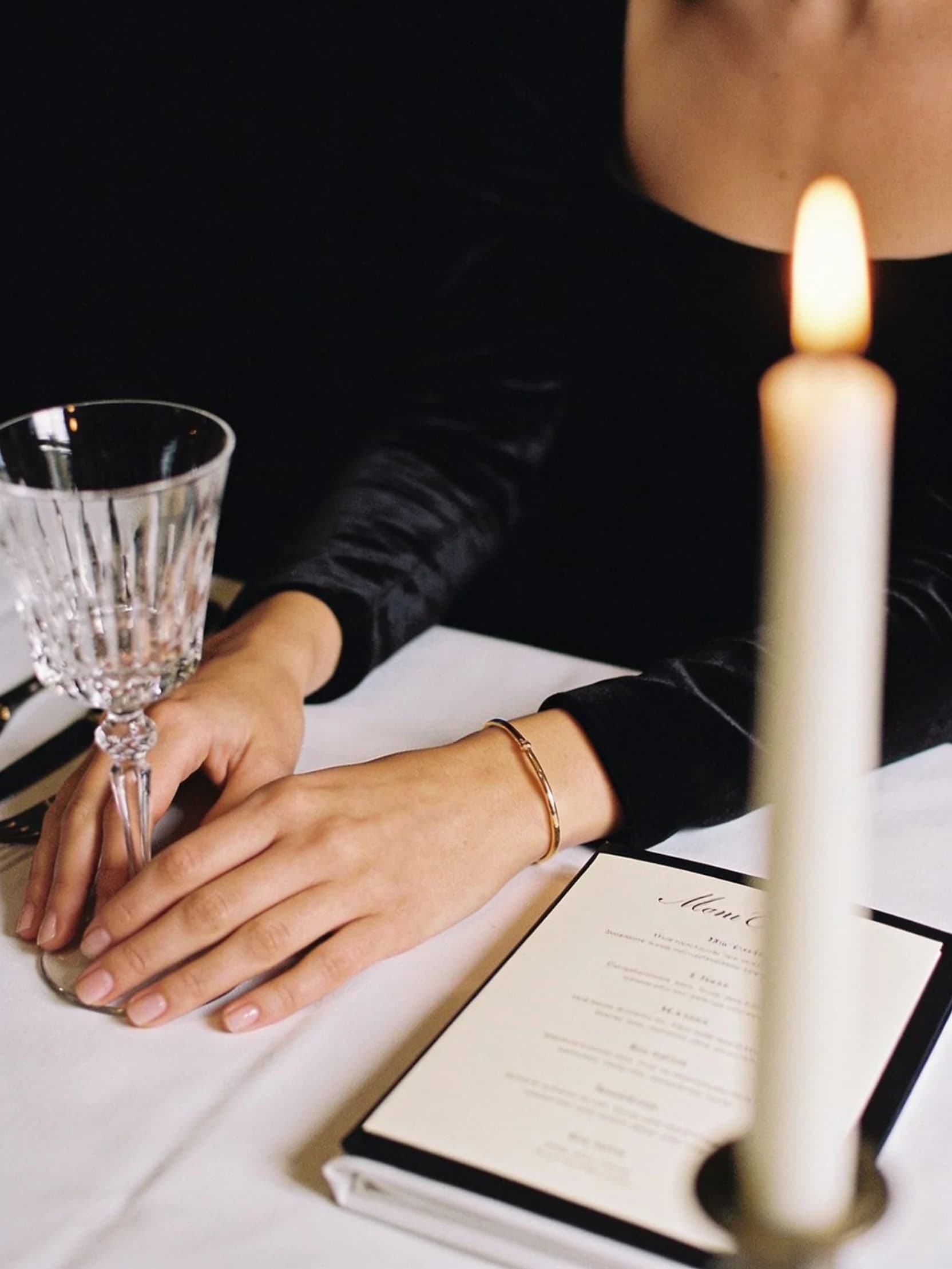 Mei-Lin's hands resting on a white linen dinner table beside a crystal glass, by candlelight.