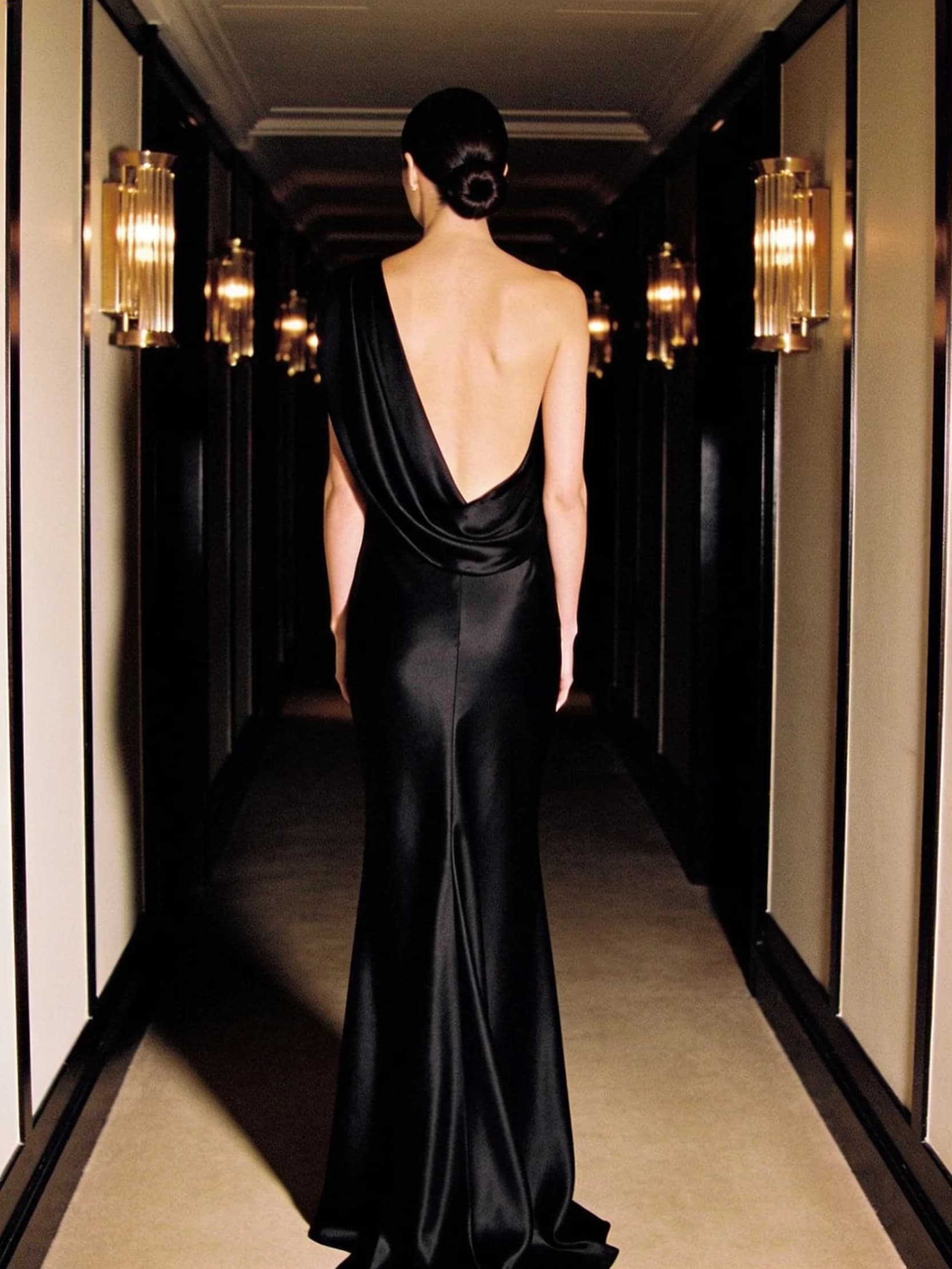 Priya, photographed from behind in a floor-length black silk gown in a lamplit hotel corridor.