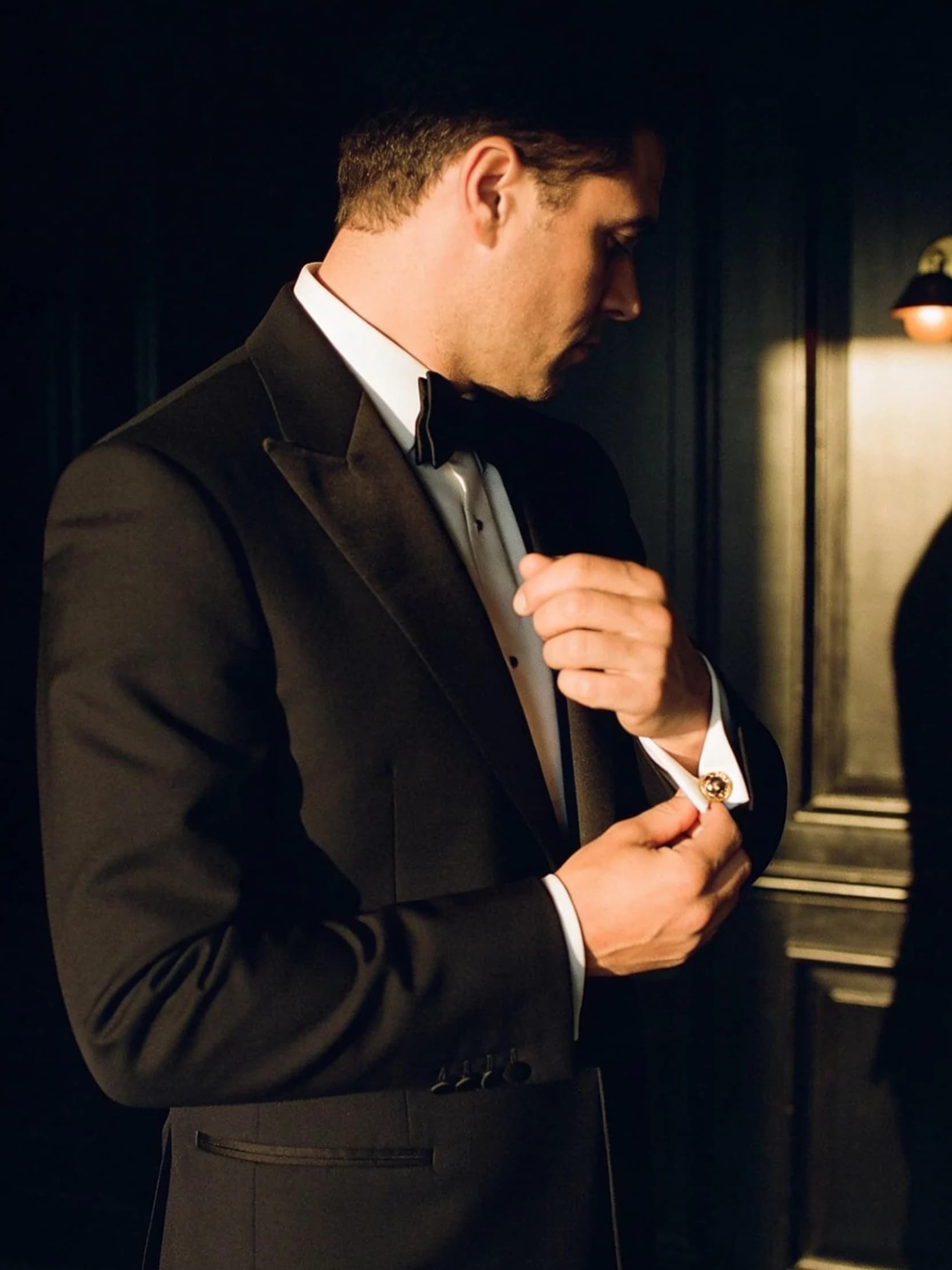 Theo in a black dinner jacket, adjusting a cuff, photographed from behind in a panelled room.